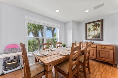 Sunlit coastal dining room with rustic wooden table set with bowls and glassware, ladder-back chairs, a high chair by the window, sideboard, and large window framing palm trees and ocean glimpses.