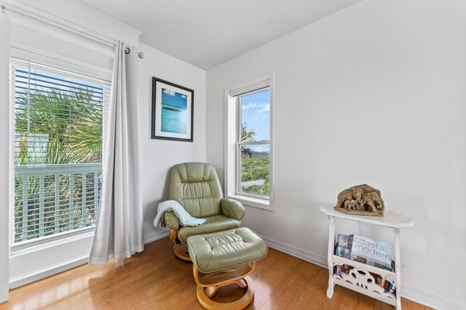Sunlit coastal reading nook with green leather recliner and matching ottoman on hardwood floor, palm trees and ocean glimpses through two windows, white walls with beach print, and a small white side table holding coastal magazines and driftwood sculpture.