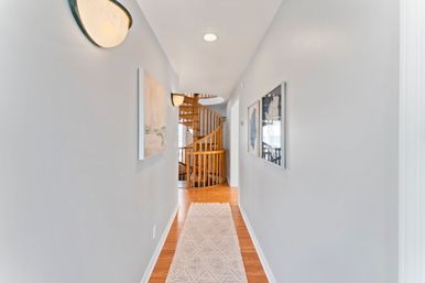 Bright interior hallway with light-gray walls, hardwood floor and woven runner, framed artwork and wall sconces leading to a natural wood spiral staircase.