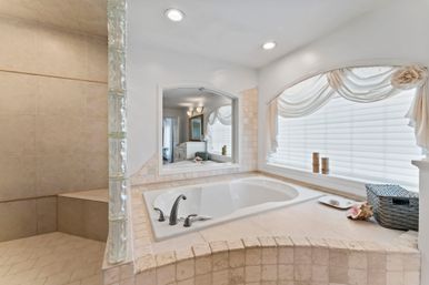 Sunlit spa-like master bathroom featuring a tiled built-in soaking tub with brushed nickel faucet, glass-block shower partition, arched window with sheer valance, and decorative basket accents.