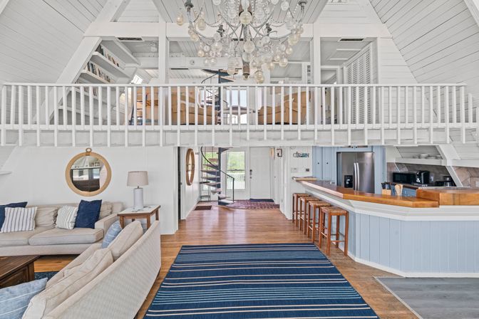 Bright coastal A-frame loft with white shiplap, open mezzanine and chandelier, spiral staircase, blue-striped rug, kitchen bar with wooden stools, and cozy beige sofas