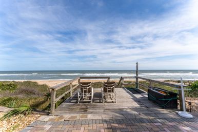 Two lounge chairs on a weathered wooden deck overlooking sandy dunes and gentle ocean waves under a bright blue sky