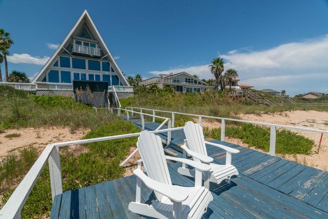Beachfront A-frame house with large windows behind a blue wooden boardwalk and two white Adirondack chairs, sandy dunes and palm trees under a bright blue sky