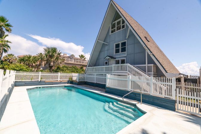 Sunny A-frame beach house with steep gabled roof, white picket railing, palm trees and a turquoise private pool.