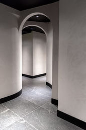 Sleek minimalist interior corridor with smooth pale plaster walls, black baseboards, repeating arched doorways and dark stone tile floor lit by recessed ceiling lights.