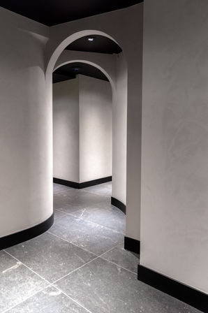 Sleek minimalist interior corridor with smooth pale plaster walls, black baseboards, repeating arched doorways and dark stone tile floor lit by recessed ceiling lights.