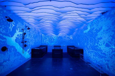 Blue-lit indoor wellness room with three black cold-plunge tubs, wall-mounted shower heads, textured illuminated walls and layered wave ceiling creating an underwater spa vibe.