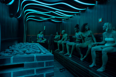 Group of people relaxing in a modern LED-lit wood-paneled spa sauna, sitting on benches near heated stones under wavy blue ceiling lights.