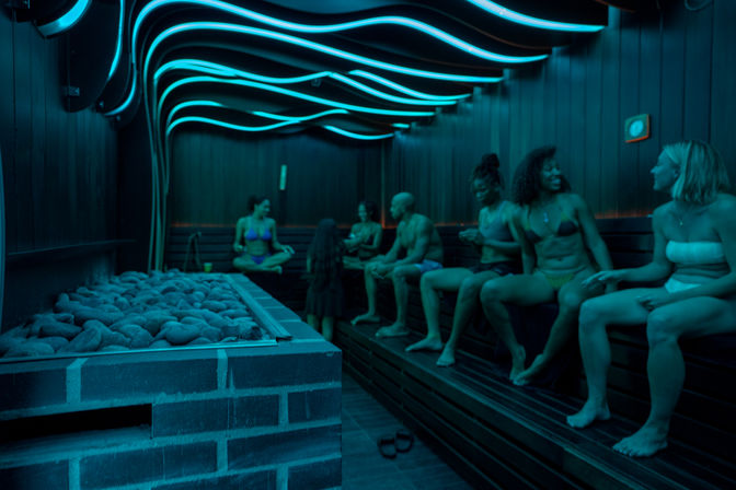 Group of people relaxing in a modern LED-lit wood-paneled spa sauna, sitting on benches near heated stones under wavy blue ceiling lights.