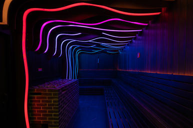 Neon-lit modern sauna interior with wavy pink, purple and blue LED ceiling lights, brick heater with stones and long wooden bench seating