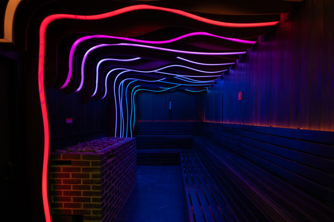 Neon-lit modern sauna interior with wavy pink, purple and blue LED ceiling lights, brick heater with stones and long wooden bench seating