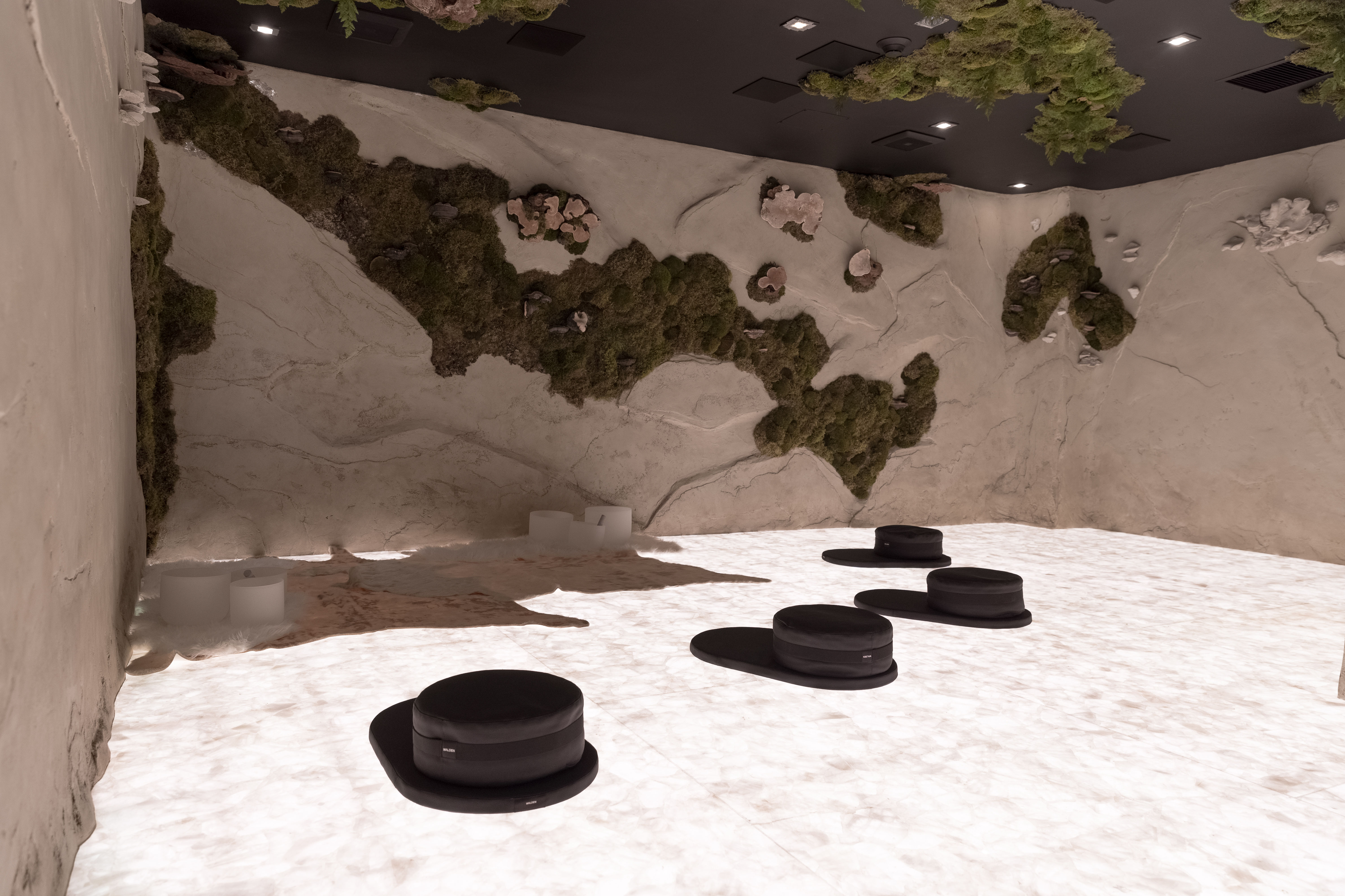 Calm modern zen meditation studio with glowing white stone floor, textured beige walls with moss wall art, ambient ceiling lights, and low round black floor cushions arranged for a spa or wellness lounge.