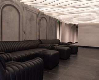Modern minimalist lounge interior with ribbed black leather sofas and ottomans along a textured arched wall, sculptural wavy ceiling lights casting warm ambient glow over dark tile floor.