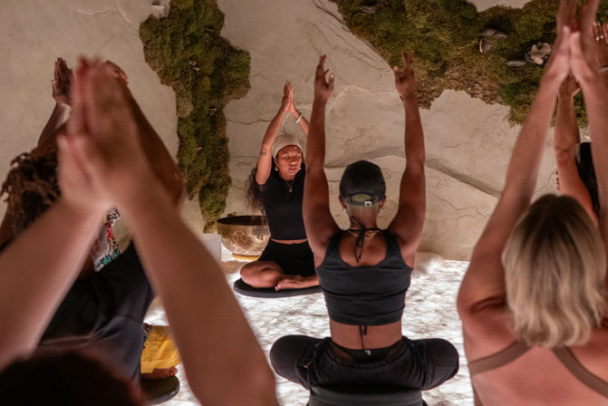 Group yoga/meditation class in a cozy wellness studio, instructor seated on a cushion with hands raised while participants mirror the pose in front of a moss-covered wall and softly glowing floor.