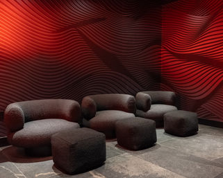 Contemporary lounge with rounded black bouclé armchairs and cube ottomans on stone tile, set against a dramatic red wavy-pattern accent wall and moody lighting.