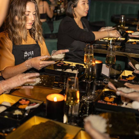 Hands-on sushi-making workshop at a candlelit dinner table — guests in aprons and gloves spreading rice on nori sheets with bento-style trays and champagne glasses.