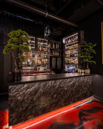 Modern upscale cocktail bar interior with a dark marble front counter, backlit shelves of liquor bottles and glassware, potted bonsai trees flanking the bar, glossy red swirled resin floor and warm ambient lighting.