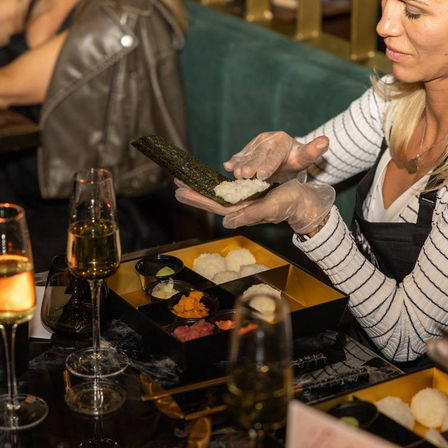 Gloved hands assembling a nori hand roll with sushi rice, bento-style tray of assorted fillings and champagne glasses on a restaurant table — sushi-making experience.