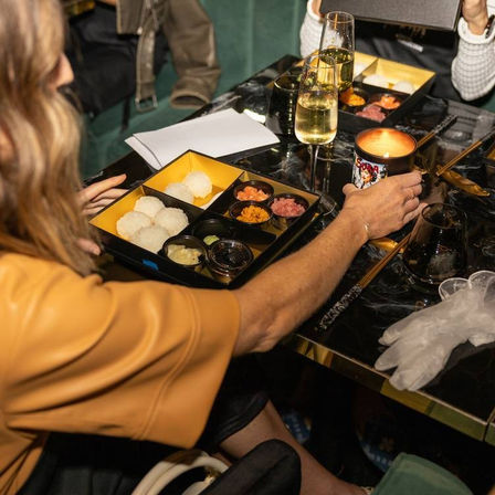 Stylish sushi bento dinner at a black marble table — rice balls, small condiment cups, chopsticks, champagne flutes and a lit candle beside diners’ hands.