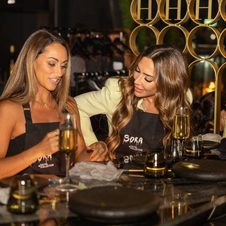 Two women in black aprons at an upscale bar table, one showing the other something on the table while champagne flutes and warm golden lights reflect on a glossy surface, cozy evening atmosphere.