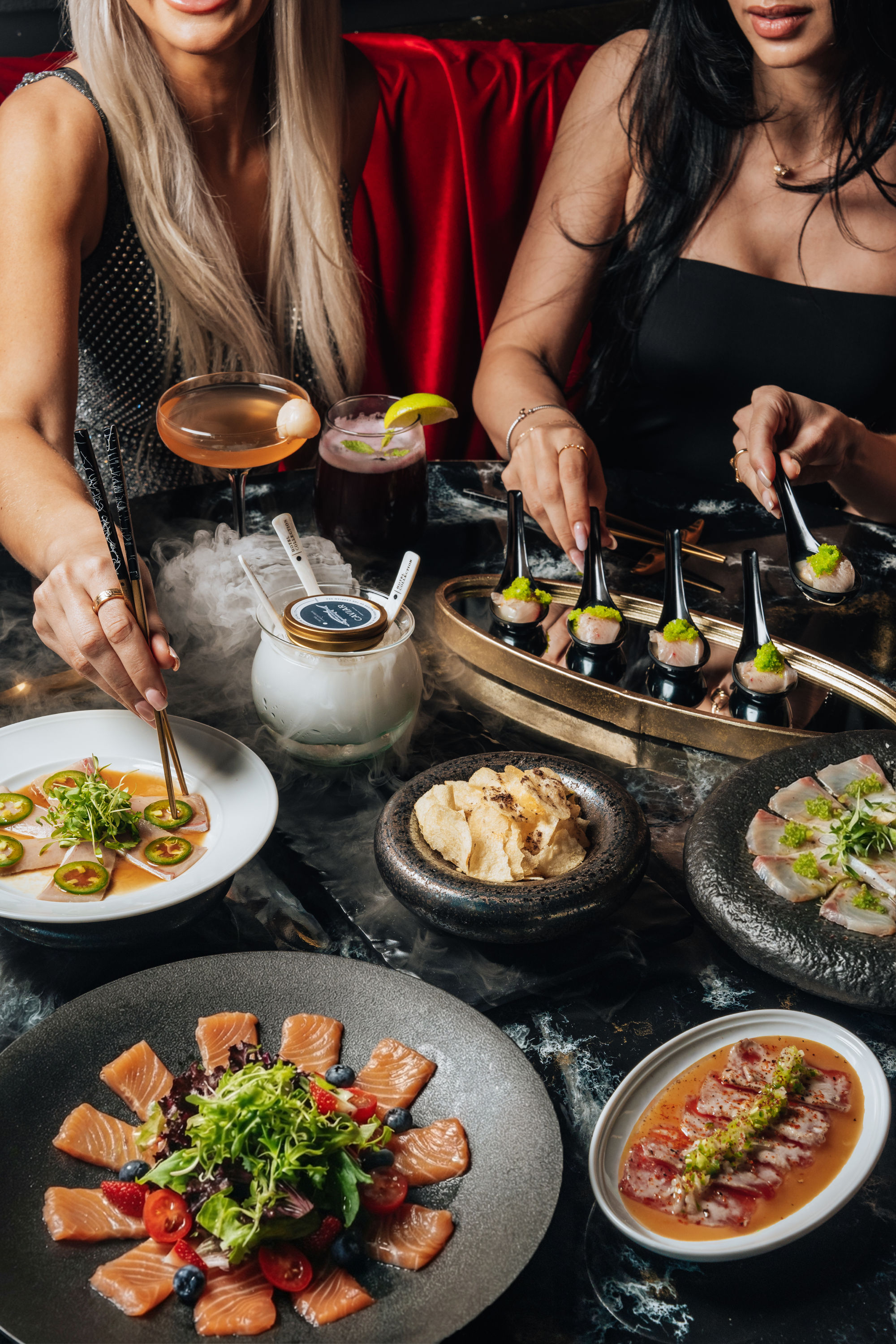 Two diners enjoying sushi and sashimi bites with cocktails and smoky dry-ice presentation on a marble table in an upscale city restaurant.