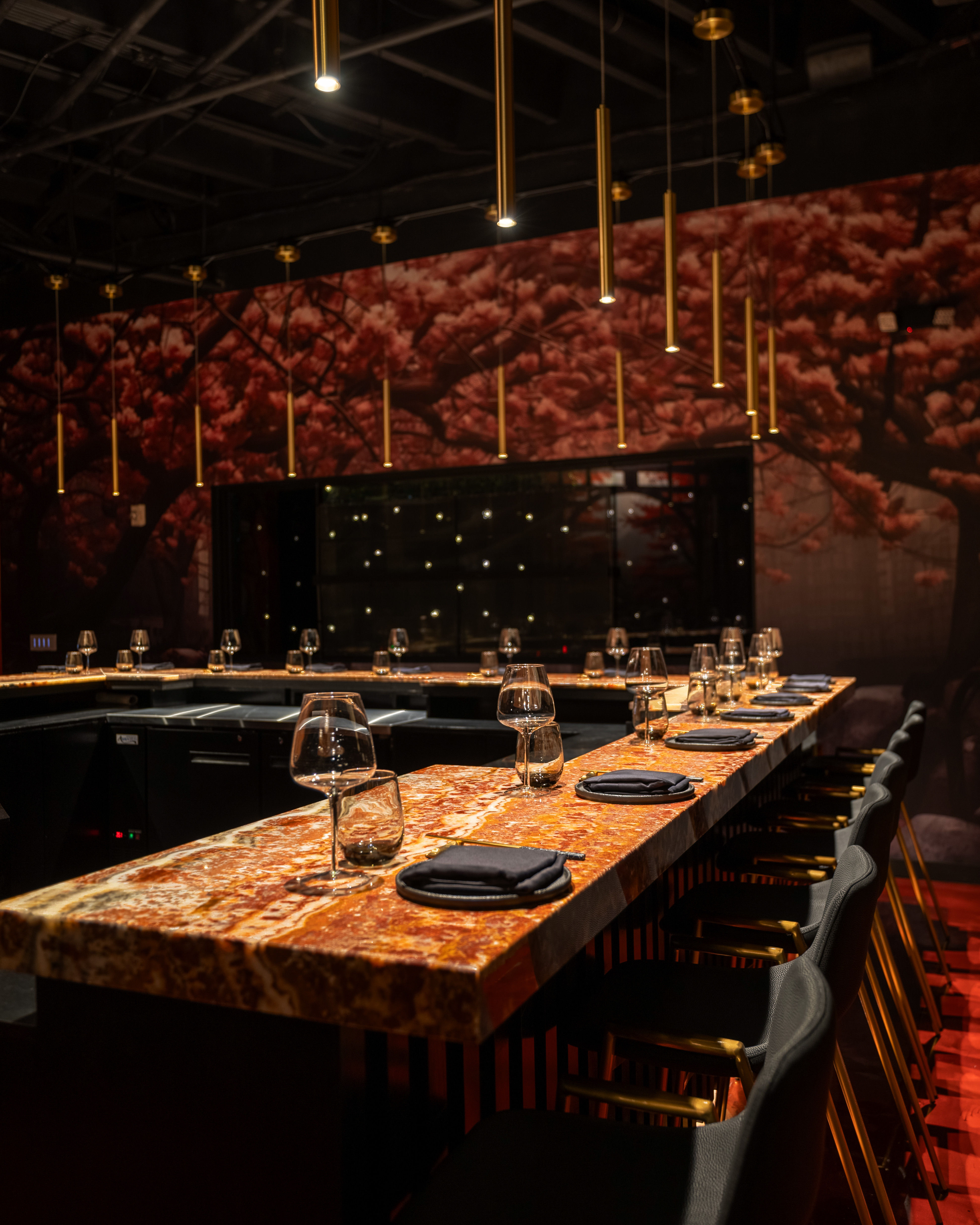 Luxurious U-shaped marble bar with amber-toned countertop, hanging brass pendant lights, black bar stools, wine glasses and napkins set against a pink cherry blossom wall mural — upscale urban restaurant interior.