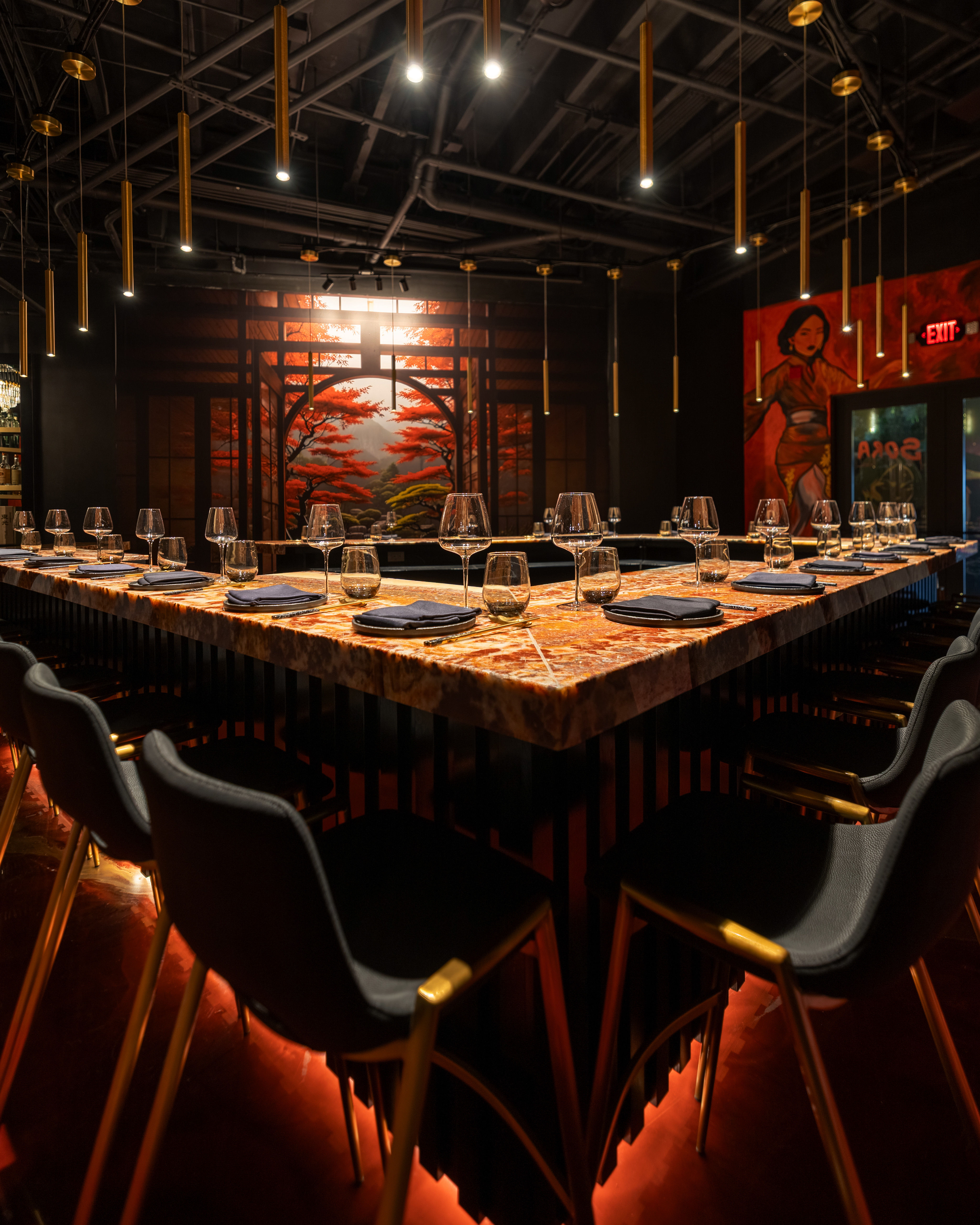 Moody Asian-inspired dining space with a U-shaped marble bar set with wine glasses and napkins, black chairs with gold legs, hanging brass pendant lights, warm red underlighting, and a large mural of red maple trees.