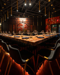 Moody Asian-inspired dining space with a U-shaped marble bar set with wine glasses and napkins, black chairs with gold legs, hanging brass pendant lights, warm red underlighting, and a large mural of red maple trees.