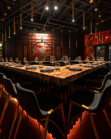 Moody Asian-inspired dining space with a U-shaped marble bar set with wine glasses and napkins, black chairs with gold legs, hanging brass pendant lights, warm red underlighting, and a large mural of red maple trees.