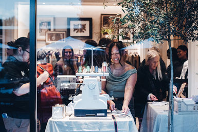 Sewing demonstration through a boutique storefront window: smiling artisan at a serger machine with colorful threads and a crowd of shoppers watching