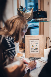 Shopper filling out cards at a boutique display table with a framed embroidered patch menu, scattered pens, a cocktail glass, and handbags on a glass shelf