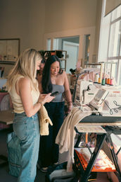 Two women smiling at a smartphone beside an embroidery machine with colorful thread spools in a sunlit sewing studio boutique