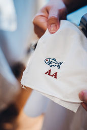 Close-up of a white embroidered handkerchief held by hands, featuring a small blue fish motif and red "AA" initials, soft indoor lighting