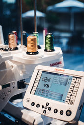 Close-up of an embroidery machine control panel with colorful thread spools and an LCD preview of a fish and "VB" monogram, sewing equipment in a craft studio