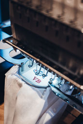 Close-up of an embroidery machine stitching a small blue motif and red "YB" monogram on white fabric in a textile studio