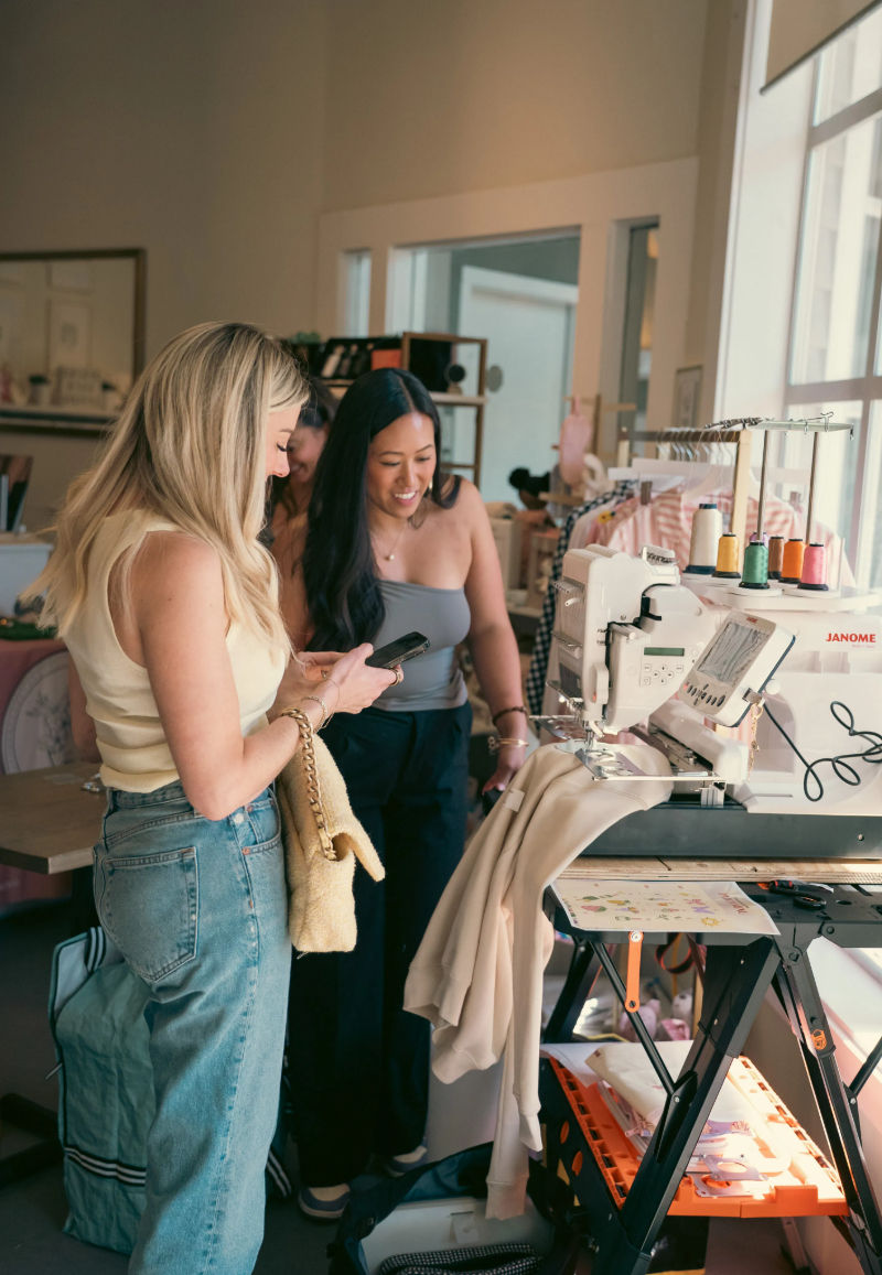 Two women smiling in a bright sewing studio, checking a phone beside a sewing/embroidery machine threaded with colorful spools, with garments on racks and fabric draped over the worktable.