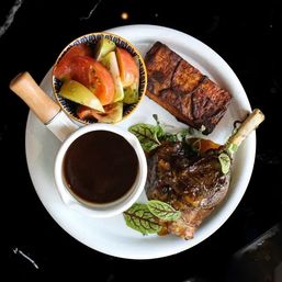 Top-down restaurant-style plate with braised lamb shank glazed in gravy, cup of dark sauce, grilled plantain and a tomato‑cucumber salad garnished with microgreens