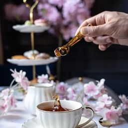 Close-up of a white porcelain teacup as golden honey drips from an amber honey dipper, splash captured; elegant afternoon tea table with pink blossoms and a two-tier pastry stand blurred in the background.