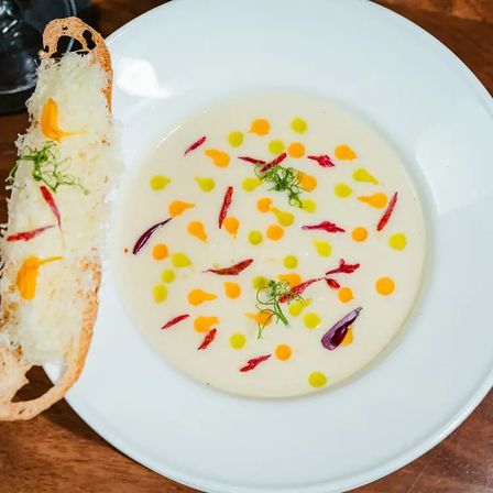 Gourmet creamy white soup in a white bowl dotted with yellow and orange oil drops, red edible petals and microgreens, served with a cheese-topped crostini on a wooden table