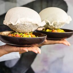 Two black ceramic bowls of colorful diced vegetable salad, each topped with a large airy white puffed bread dome, held on hands as a playful gourmet restaurant presentation.