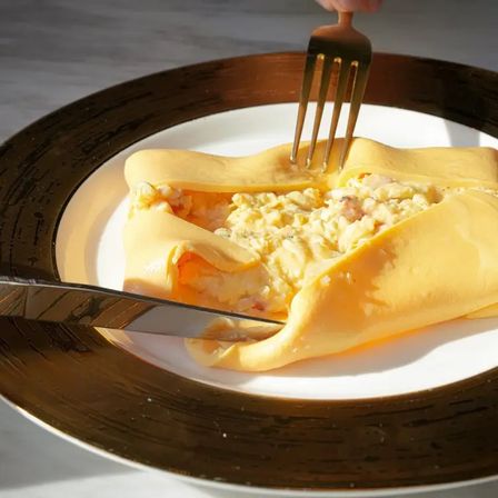 Fork and knife cutting into a folded golden omelette oozing creamy scrambled-egg filling on a white plate — gourmet breakfast/brunch dish