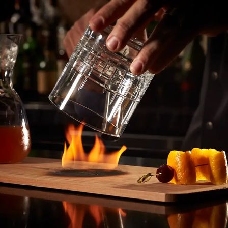 Flame-smoked cocktail scene: hands place a crystal whiskey glass over a small fire on a wooden board next to orange peel and cherry garnish in a dim craft cocktail bar.