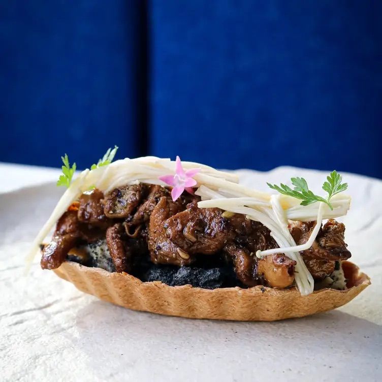 Savory mini tartlet with braised soy-glazed meat, enoki mushrooms, dark sesame-like filling, edible pink flower and microgreens against a blue backdrop.