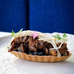 Savory mini tartlet with braised soy-glazed meat, enoki mushrooms, dark sesame-like filling, edible pink flower and microgreens against a blue backdrop.