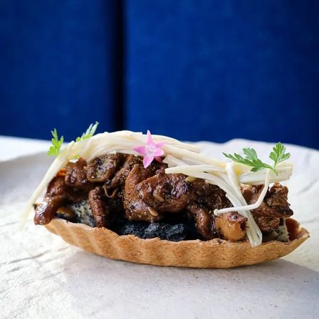 Savory mini tartlet with braised soy-glazed meat, enoki mushrooms, dark sesame-like filling, edible pink flower and microgreens against a blue backdrop.