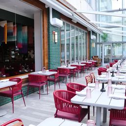 Sunlit urban al fresco restaurant patio with rows of red woven chairs and white tables along a green tiled wall under a glass atrium.