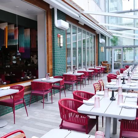 Sunlit urban al fresco restaurant patio with rows of red woven chairs and white tables along a green tiled wall under a glass atrium.