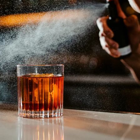 Amber whiskey cocktail in a faceted rocks glass with ice and citrus peel as a bartender mists an aromatic spritz over it at a dim bar