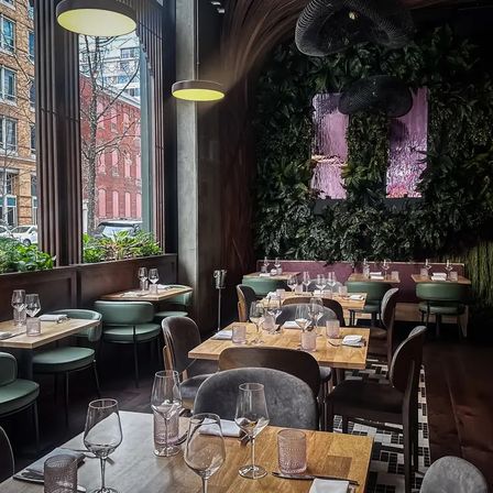 Cozy modern downtown restaurant interior with wood tables set with wine glasses, mint-green banquettes, floor-to-ceiling windows overlooking brick buildings, lush living-plant wall and pendant lights