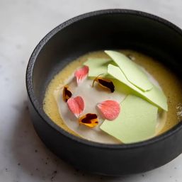Fine-dining dessert in a black ceramic bowl with silky yellow custard, white cream, pale green meringue shards and pink edible flower petals.